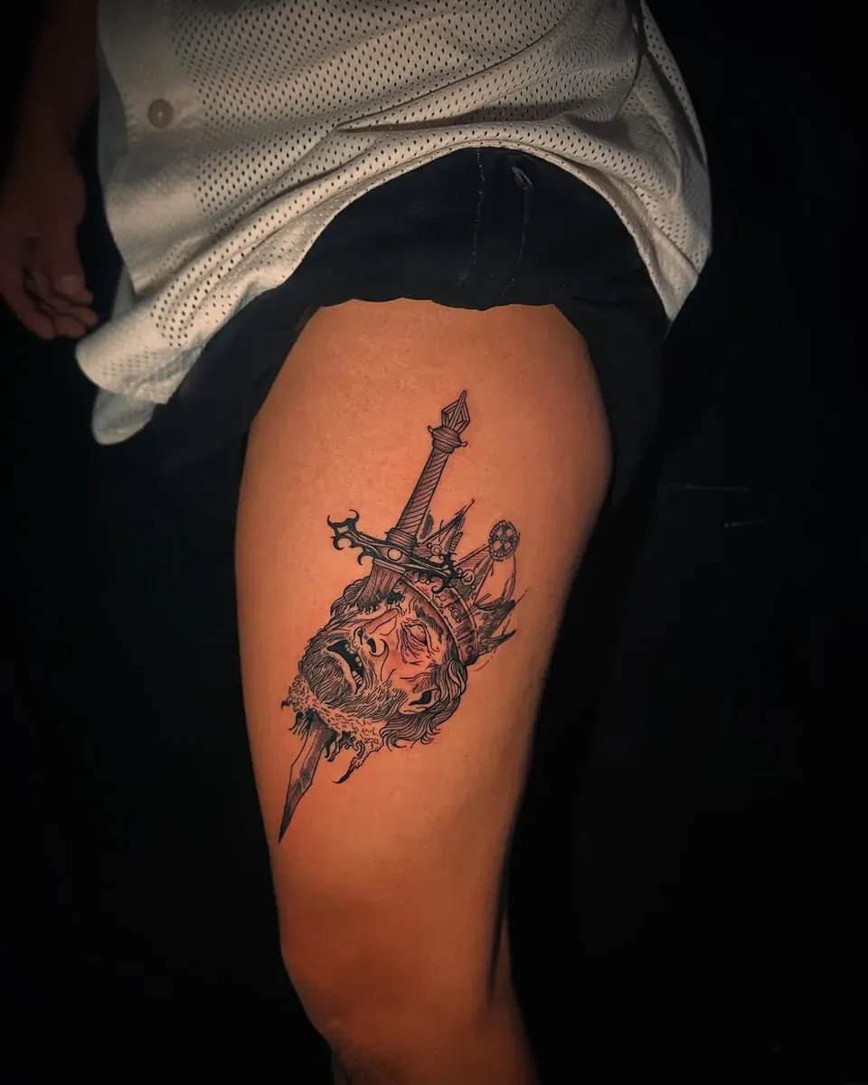 Tatuaje en black and grey de una daga con un cráneo y una corona, en el muslo